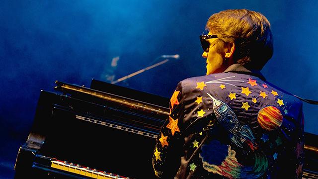 a man dressed as Elton John is seen from behind and sitting at a piano